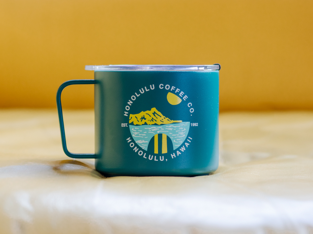 Front of a blue stainless steel mug, image showing Honolulu Coffee co, est 1992 with a drawing of a surfboard in the water. Diamond Head in the background.